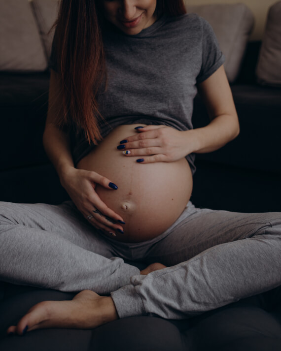 Woman in gray sweats sitting cross legged holding her exposed pregnant stomach